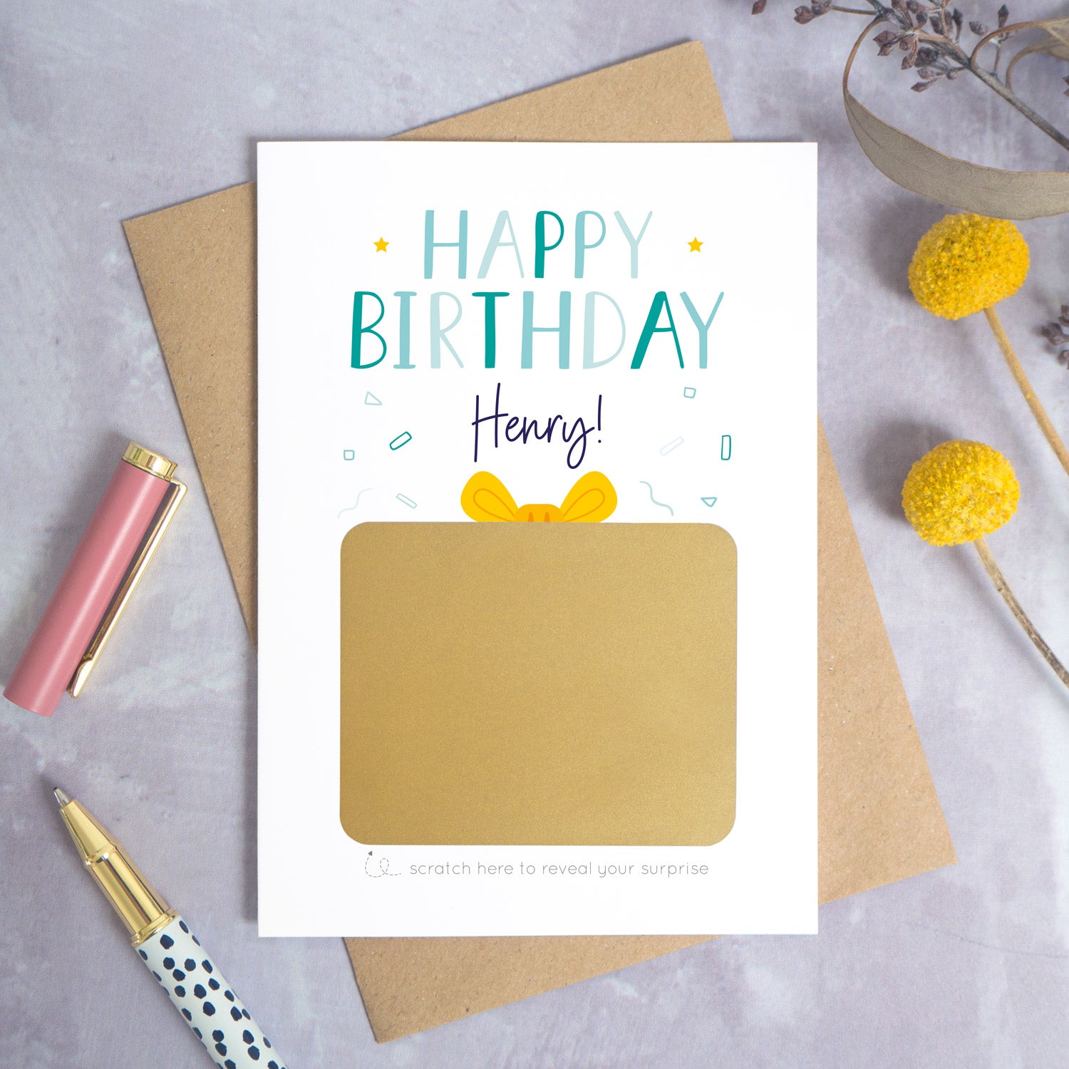 A personalised happy birthday scratch card that has been photographed flat lay style on a grey concrete style background surrounded with foliage and a pen. The card itself shows how it will arrive and looks when it is completely unscratched.