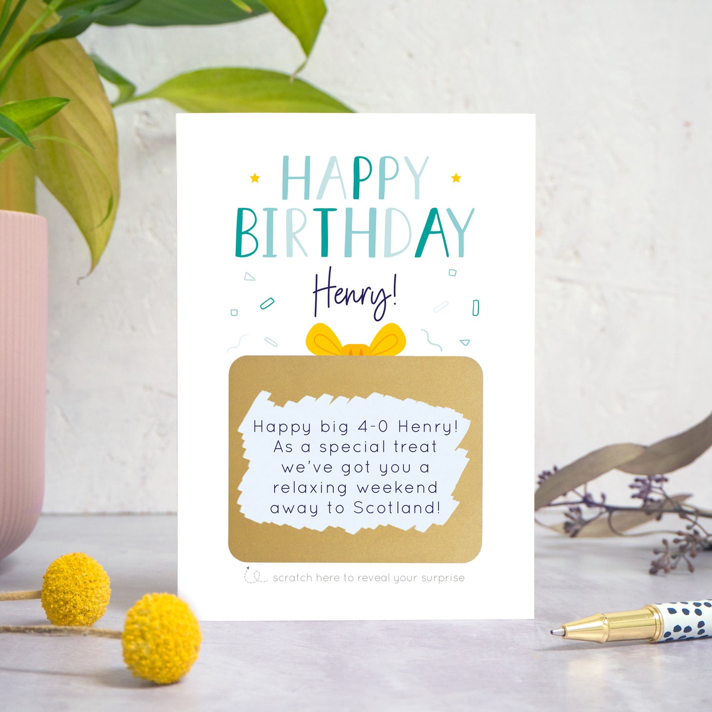 A personalised happy birthday scratch card in blue that has been photographed on a white and grey background with foliage and a pen in both the foreground and background. The card features the recipients name and a scratch panel that has been scratched off revealing a personalised message.