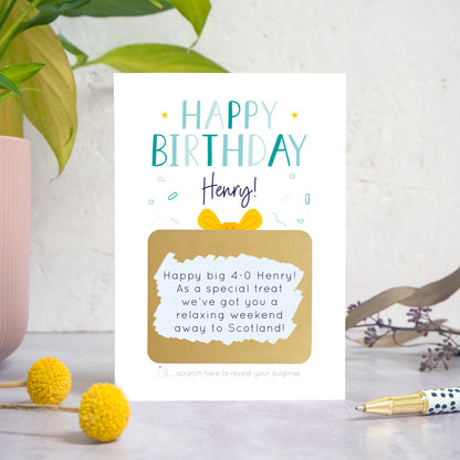 A personalised happy birthday scratch card in blue that has been photographed on a white and grey background with foliage and a pen in both the foreground and background. The card features the recipients name and a scratch panel that has been scratched off revealing a personalised message.
