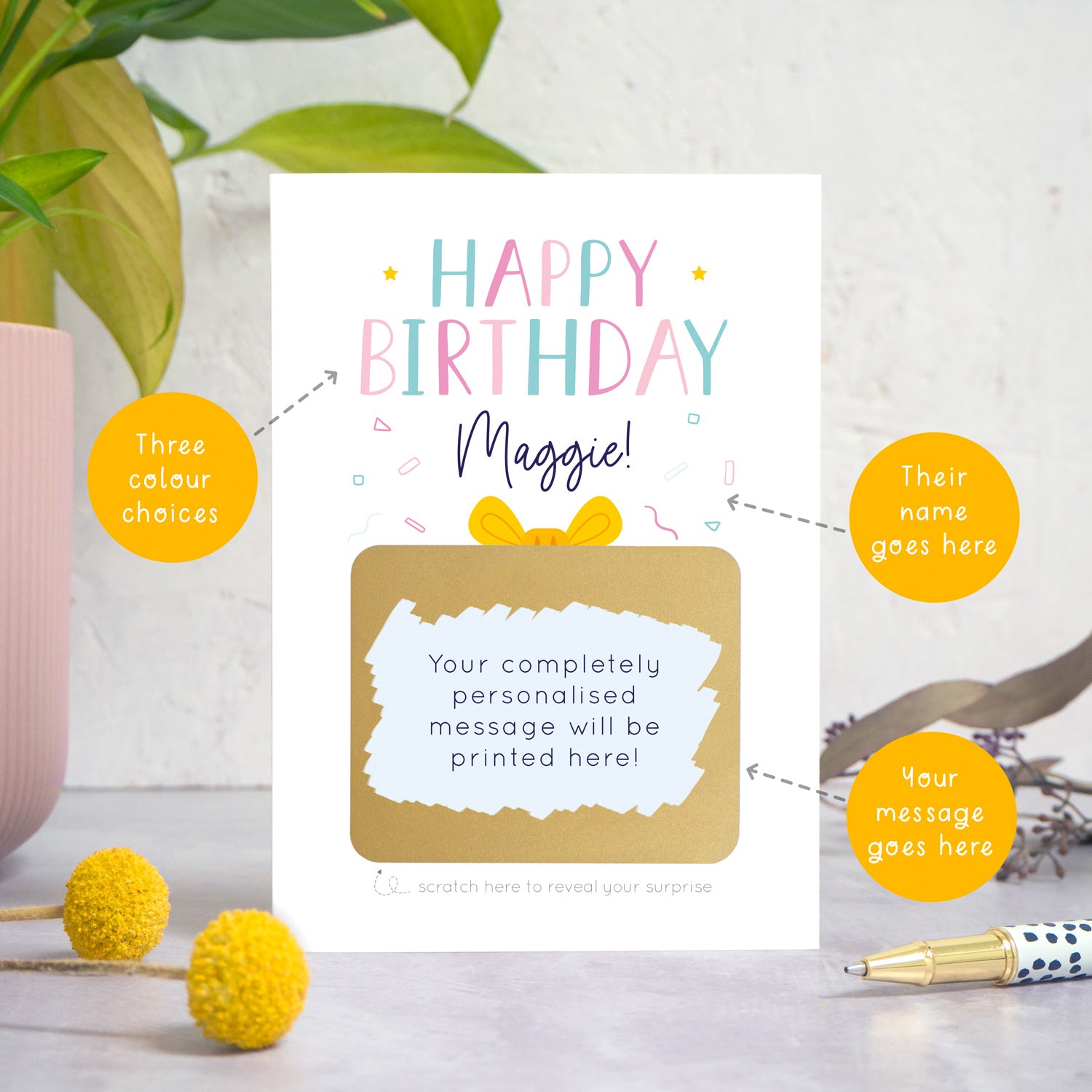 A personalised happy birthday scratch card in pink that has been photographed on a white and grey background with foliage and a pen in both the foreground and background. There are also orange circles laid over the top of this image with arrows pointing to the areas that can be customised. In this instance they point to the name, the colour choices and the personalised scratch off message.