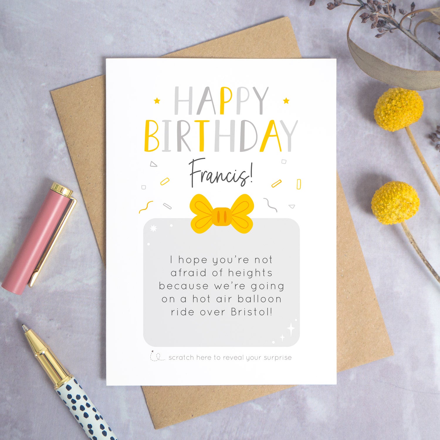 A personalised happy birthday scratch card that has been photographed flat lay style on a grey concrete style background surrounded with foliage and a pen. The card itself shows how it would look once the gold panel has been completely removed. 