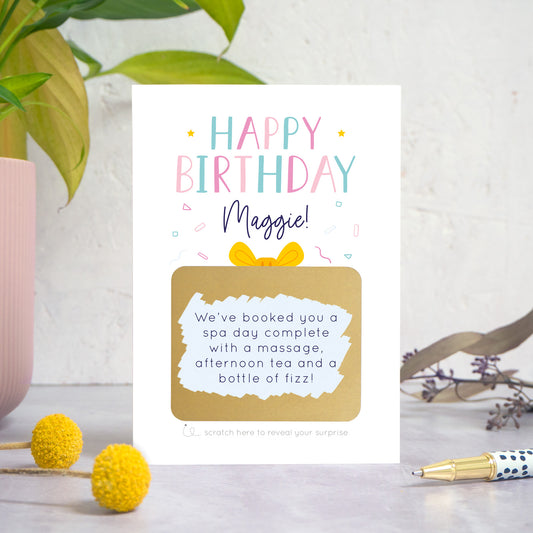 A personalised happy birthday scratch card in pink that has been photographed on a white and grey background with foliage and a pen in both the foreground and background. The card features the recipients name and a scratch panel that has been scratched off revealing a personalised message.