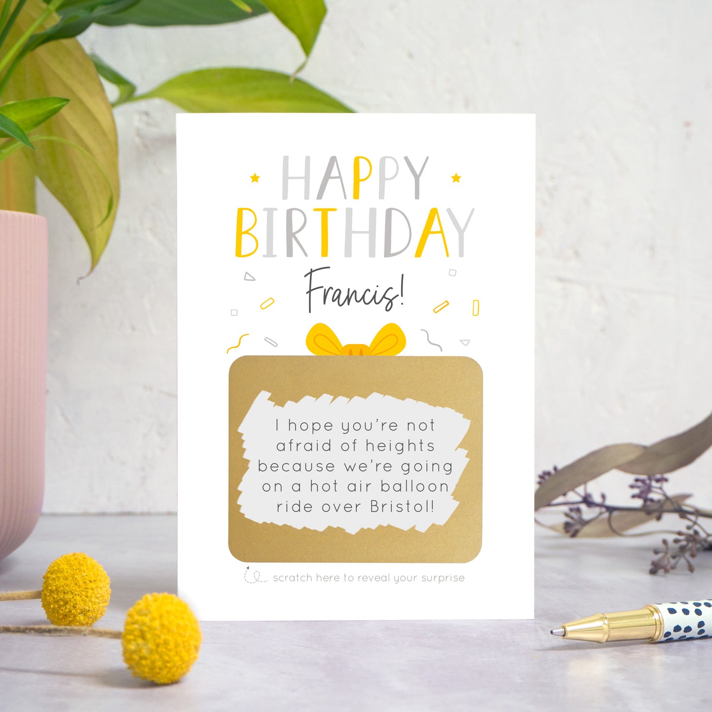 A personalised happy birthday scratch card in yellow and grey that has been photographed on a white and grey background with foliage and a pen in both the foreground and background. The card features the recipients name and a scratch panel that has been scratched off revealing a personalised message.