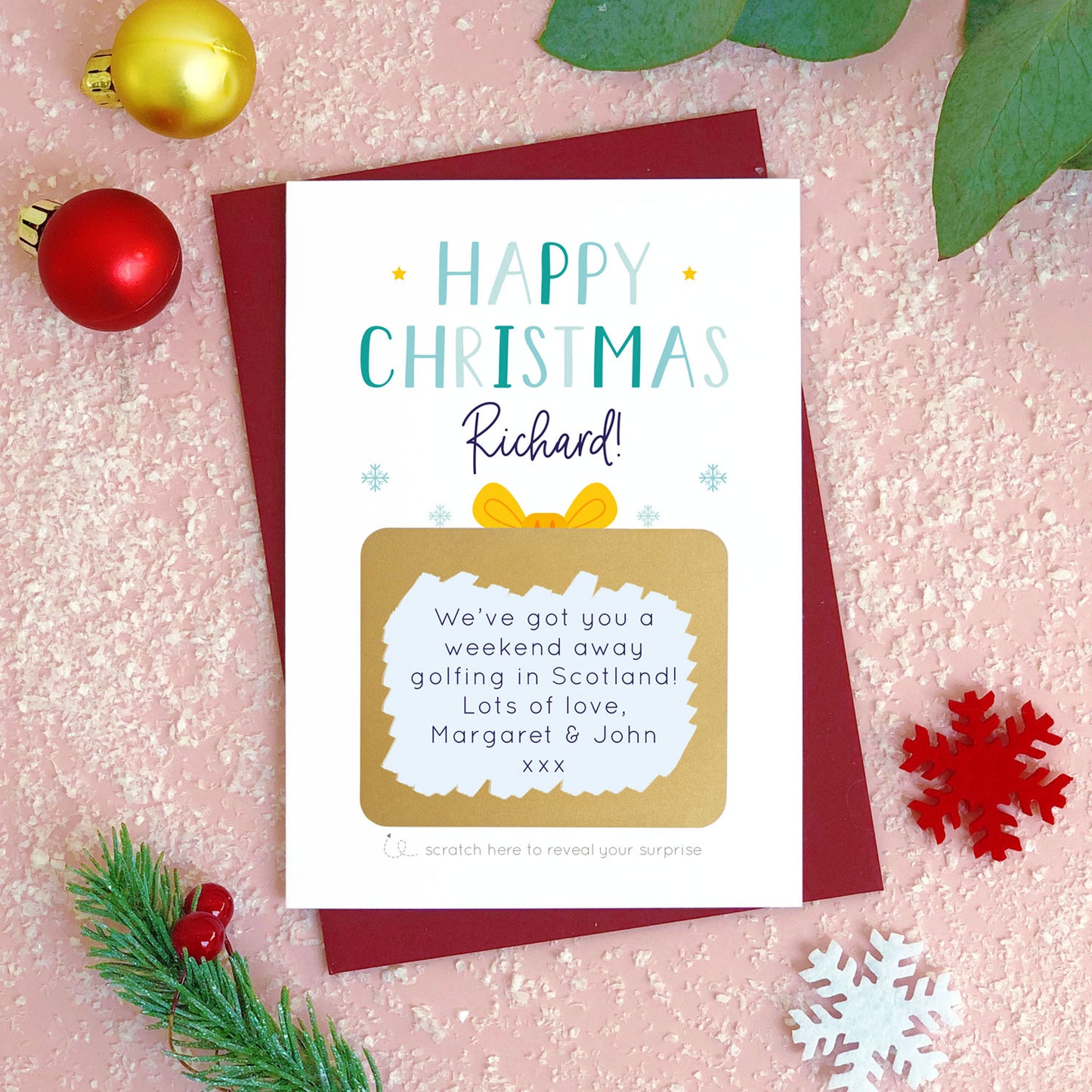 A personalised present christmas scratch card photographed flat lying on a red wine coloured envelope on a pink surface surrounded by fake snow, baubles and foliage. The gold panel has been scratched away to reveal a personalised message and gift reveal. This is the blue version of the card.