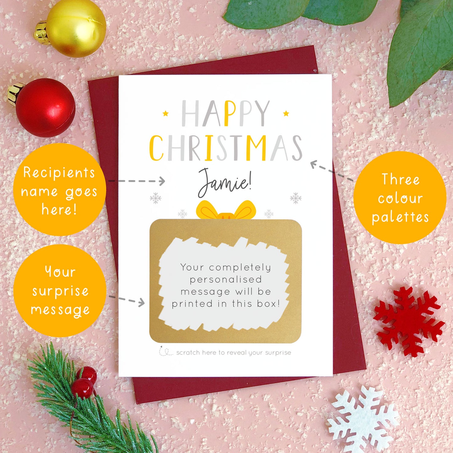 A personalised present Christmas scratch card photographed flat lying on a red wine coloured envelope. The orange circles demonstrate which areas of the card can be personalised. Here they point to the colour options, message and name for the front of the card.