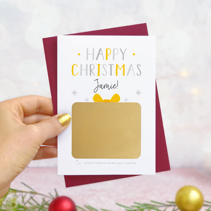 A personalised present christmas scratch card photographed being held by a hand to the left and foliage and baubles in the bottom of the frame. The card is against its ‘red wine’ colour envelope and the scratch off panel has not yet been scratched off! This is the grey & yellow version of the card.