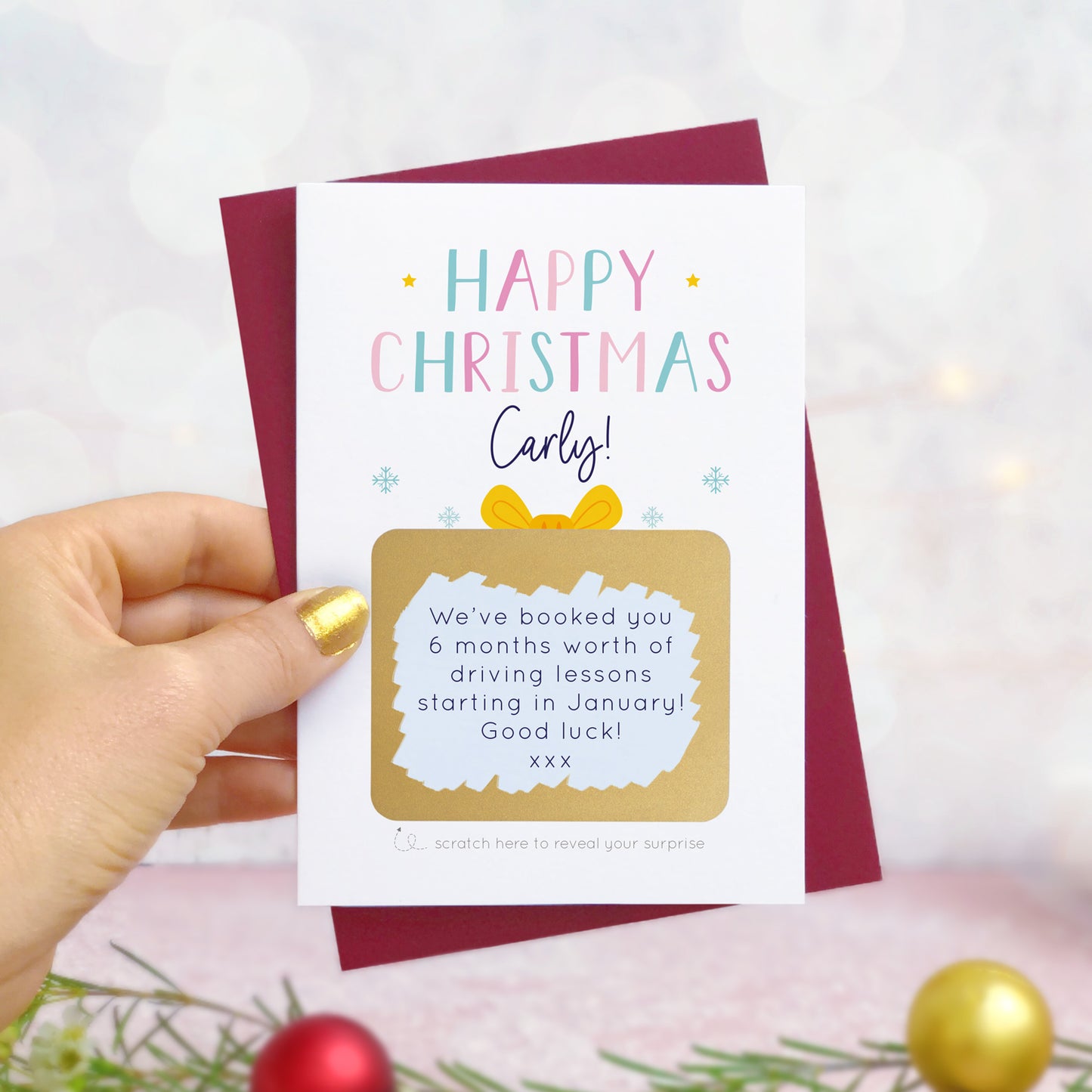 A personalised present christmas scratch card photographed being held by a hand to the left and foliage and baubles in the bottom of the frame. The card is against its ‘red wine’ colour envelope and the scratch off panel has been scratched off to reveal the personalised message! This is the pink version of the card.
