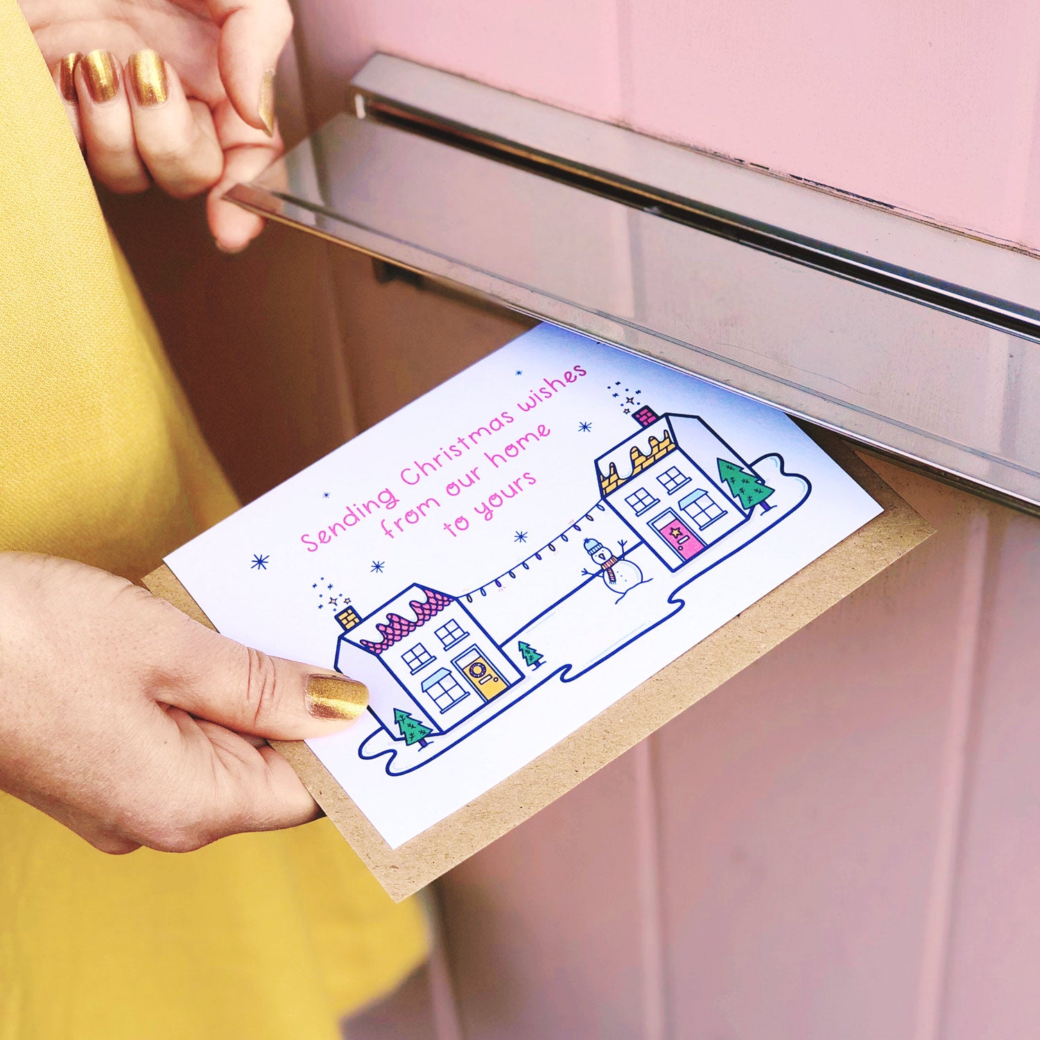 Joanne is pushing the Christmas wishes card featuring houses, snow and a snowman through the letter box of a pink door and wearing a yellow skirt.