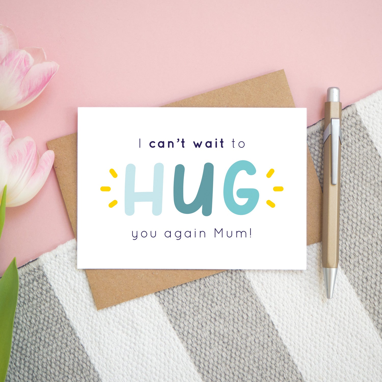 This is the personalised option of the 'I can't wait to hug you again' card with the sample name of 'Mum'. There is a pen to the right for scale and has been photographed on a pink, and grey & white striped background.