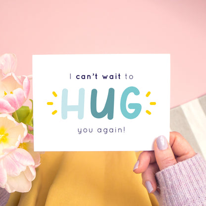 The non personalised, 'I can't wait to hug you again' card in blue. It has been shot on a pink background and has tulips to the left of the photo and is being held in the right.