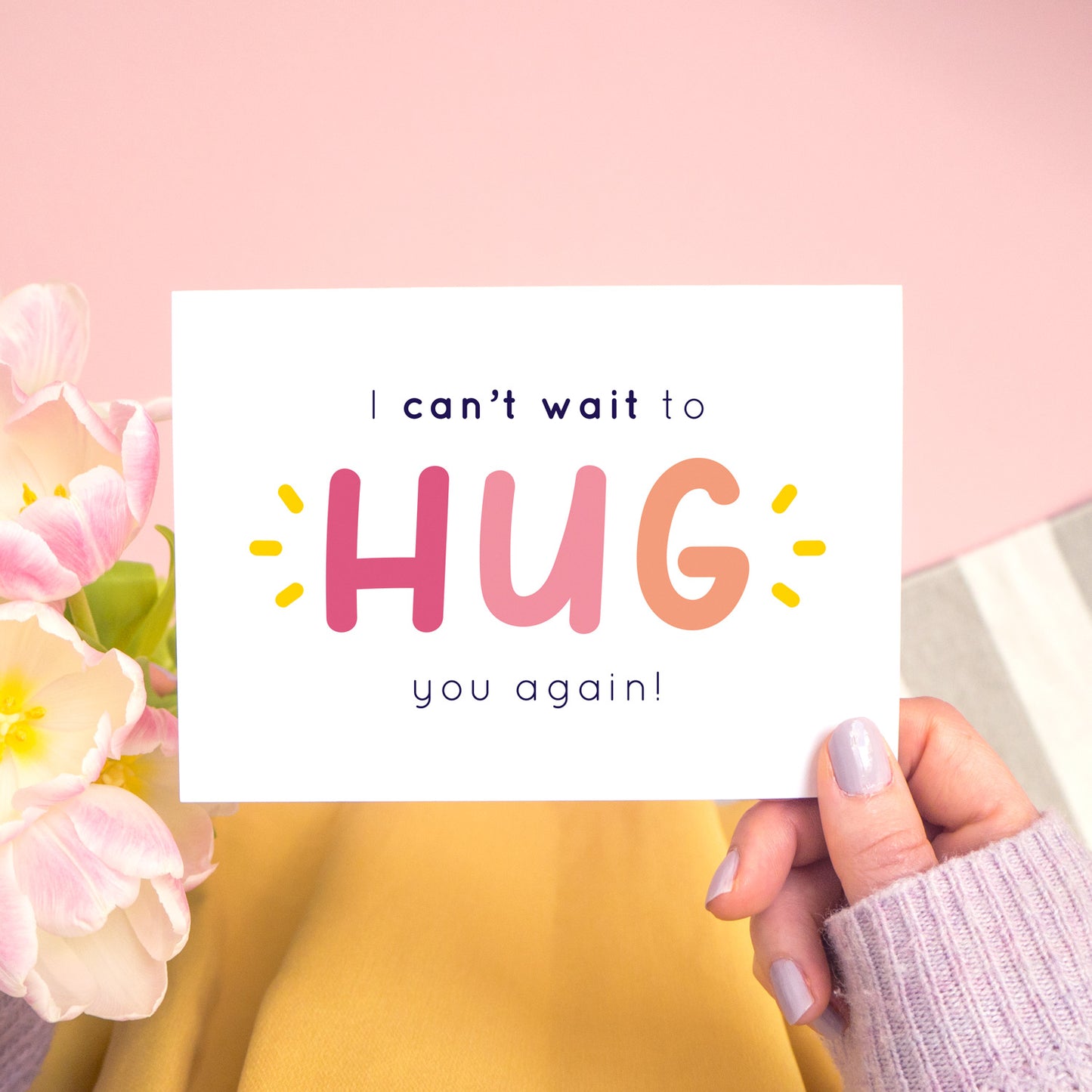 The non personalised, 'I can't wait to hug you again' card in pink. It has been shot on a pink background and has tulips to the left of the photo and is being held in the right.