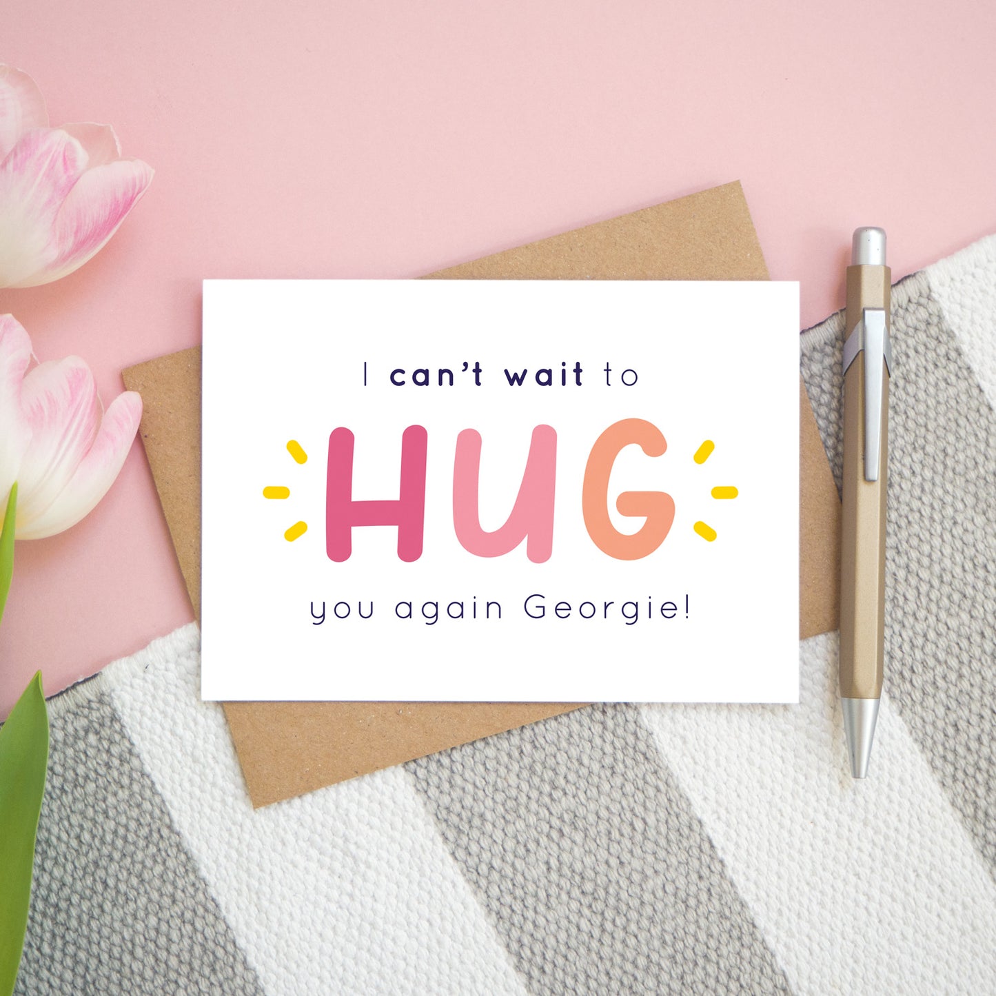 This is the personalised option of the 'I can't wait to hug you again' card with the sample name of 'Georgie'. There is a pen to the right for scale and has been photographed on a pink, and grey & white striped background.