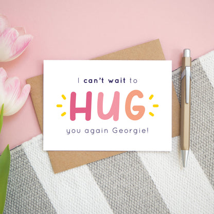 This is the personalised option of the 'I can't wait to hug you again' card with the sample name of 'Georgie'. There is a pen to the right for scale and has been photographed on a pink, and grey & white striped background.