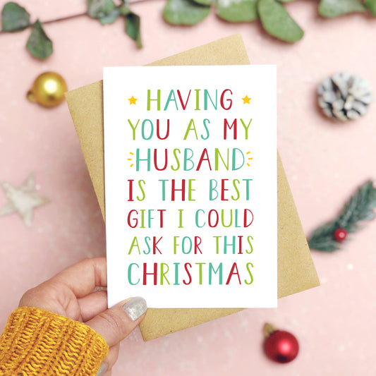 A 'best gift' husband Christmas card held over a pink background by a hand in a mustard knit jumper with foliage and baubles in the background. The writing on the card is red and green.