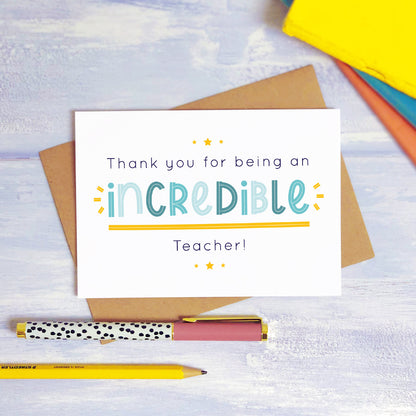 An incredible teacher thank you card lying on top of a kraft brown envelope, flat lay style on a blue textured background with colourful text books, a pen and a pencil. This teacher card features the blue text colour option.