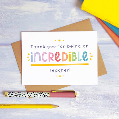 An incredible teacher thank you card lying on top of a kraft brown envelope, flat lay style on a blue textured background with colourful text books, a pen and a pencil. This teacher card features the rainbow text colour option.