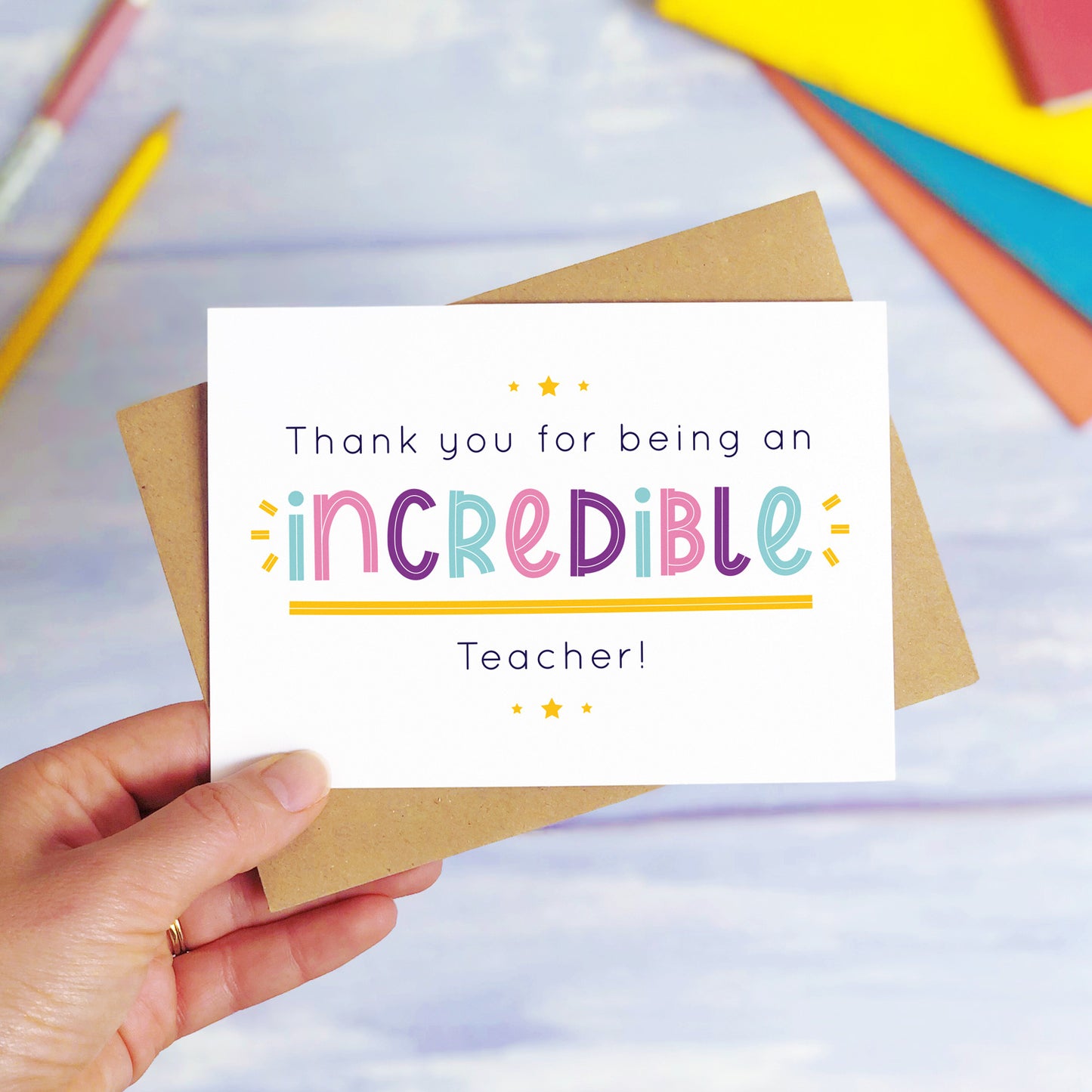 An incredible teacher thank you card being held over a blue background with colourful text books and two pens. The card is sat on top of its kraft brown envelope. This is the teacher card with the pink, purple and blue text colour option.