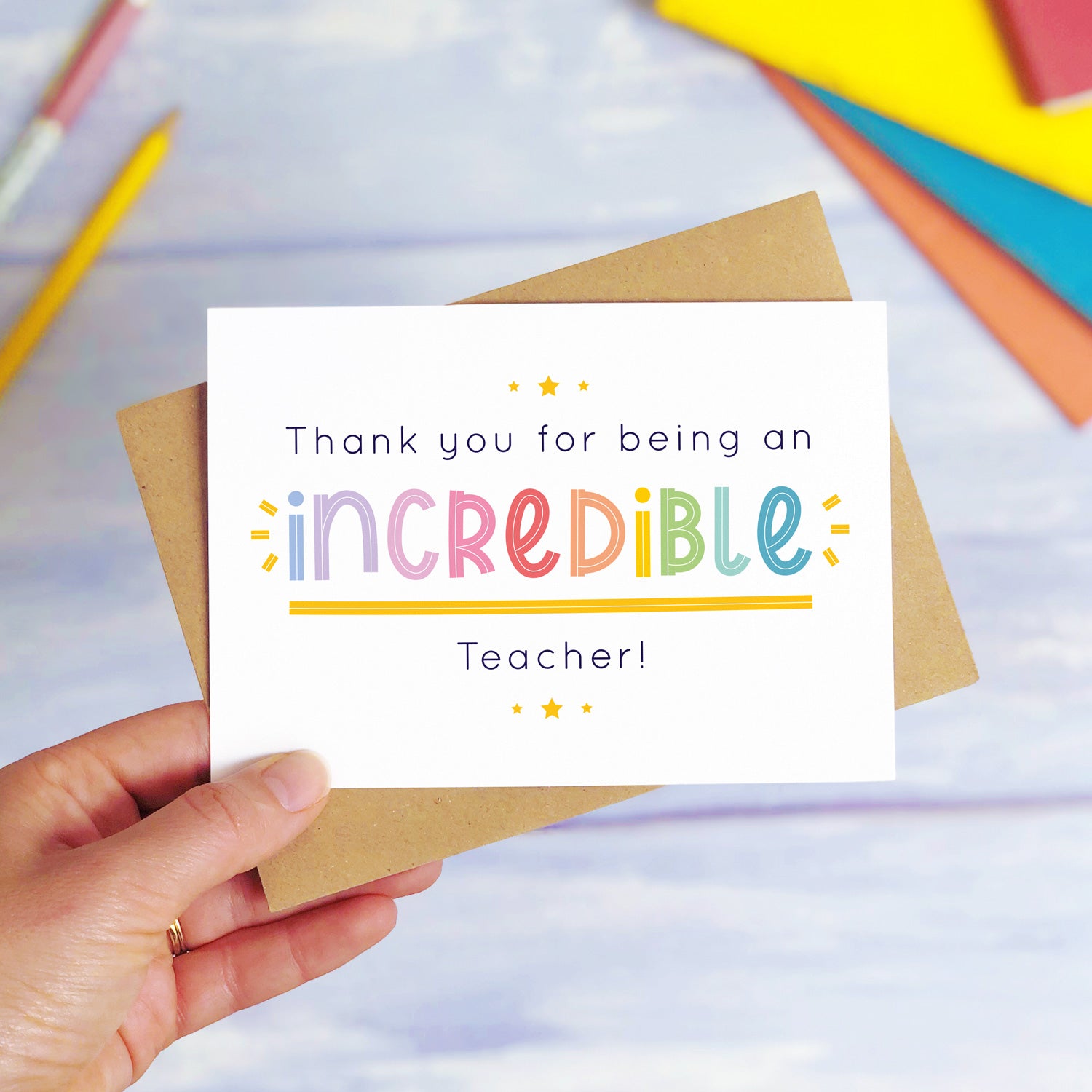 An incredible teacher thank you card being held over a blue background with colourful text books and two pens. The card is sat on top of its kraft brown envelope. This is the teacher card with the rainbow text colour option.