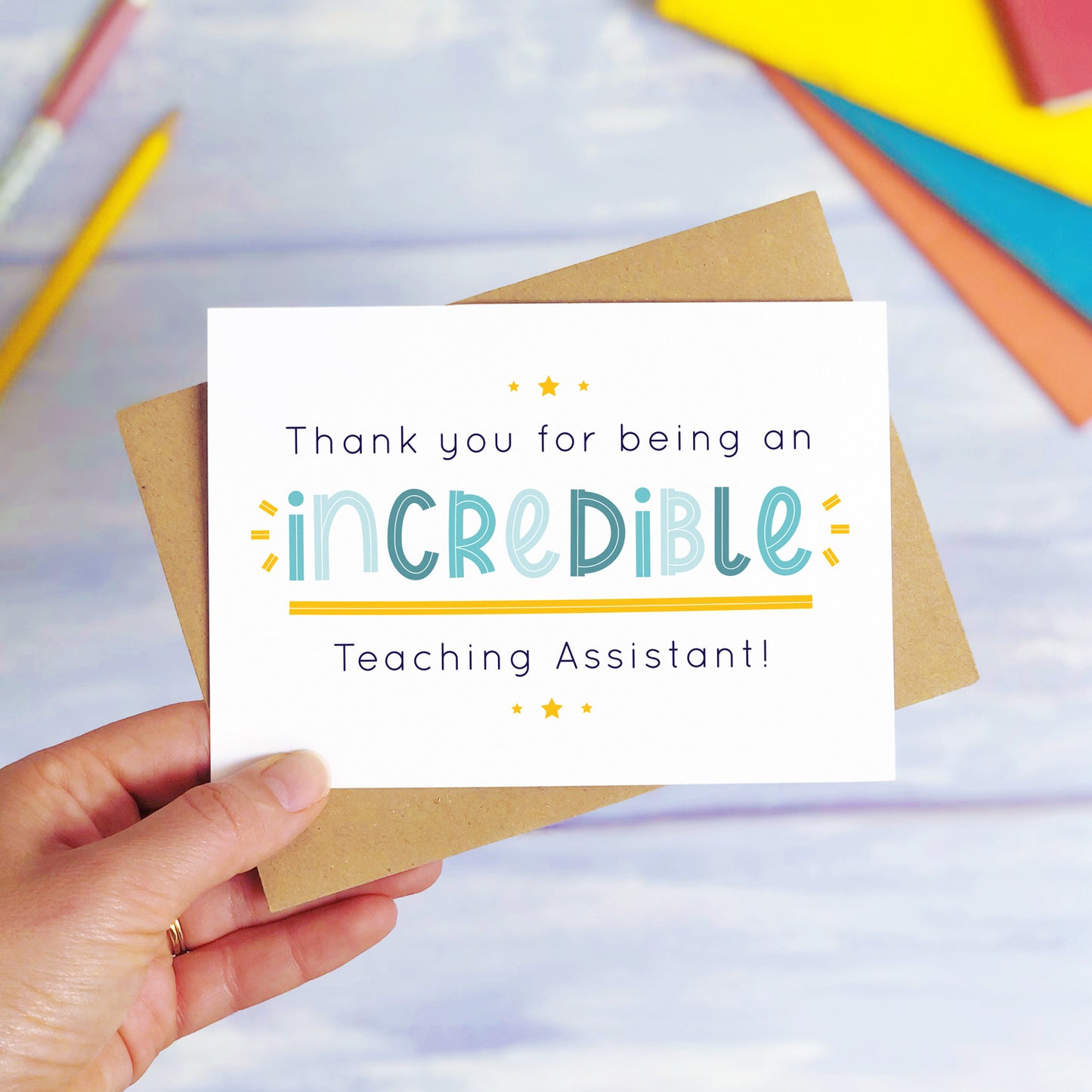 An incredible teaching assistant thank you card being held over a blue background with colourful text books and two pens. The card is sat on top of its kraft brown envelope. This is the teaching assistant card with the blue text colour option.