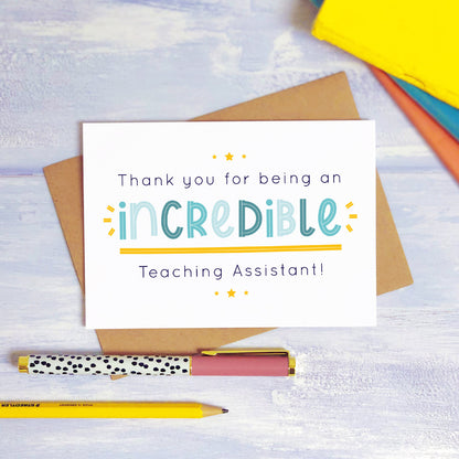 An incredible teaching assistant thank you card lying on top of a kraft brown envelope, flat lay style on a blue textured background with colourful text books, a pen and a pencil. This teaching assistant card features the blue text colour option.