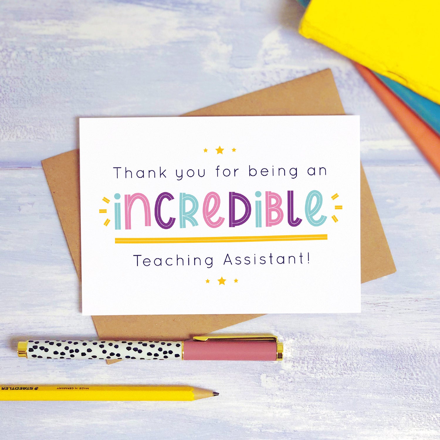 An incredible teaching assistant thank you card lying on top of a kraft brown envelope, flat lay style on a blue textured background with colourful text books, a pen and a pencil. This teaching assistant card features the pink, purple and blue text colour option.