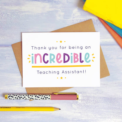 An incredible teaching assistant thank you card lying on top of a kraft brown envelope, flat lay style on a blue textured background with colourful text books, a pen and a pencil. This teaching assistant card features the pink, purple and blue text colour option.