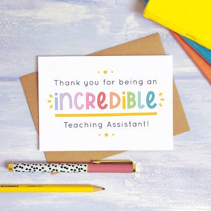 An incredible teaching assistant thank you card lying on top of a kraft brown envelope, flat lay style on a blue textured background with colourful text books, a pen and a pencil. This teaching assistant card features the rainbow text colour option.
