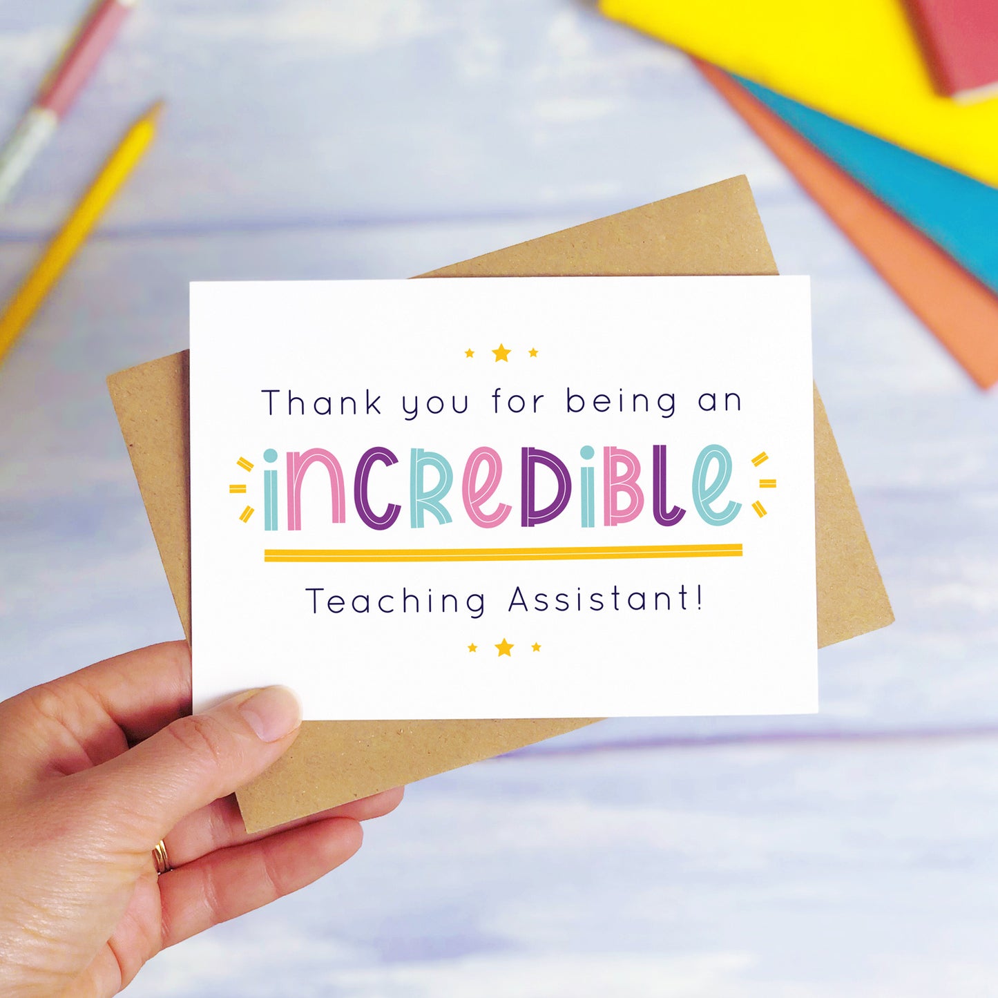 An incredible teaching assistant thank you card being held over a blue background with colourful text books and two pens. The card is sat on top of its kraft brown envelope. This is the teaching assistant card with the pink, purple and blue text colour option.