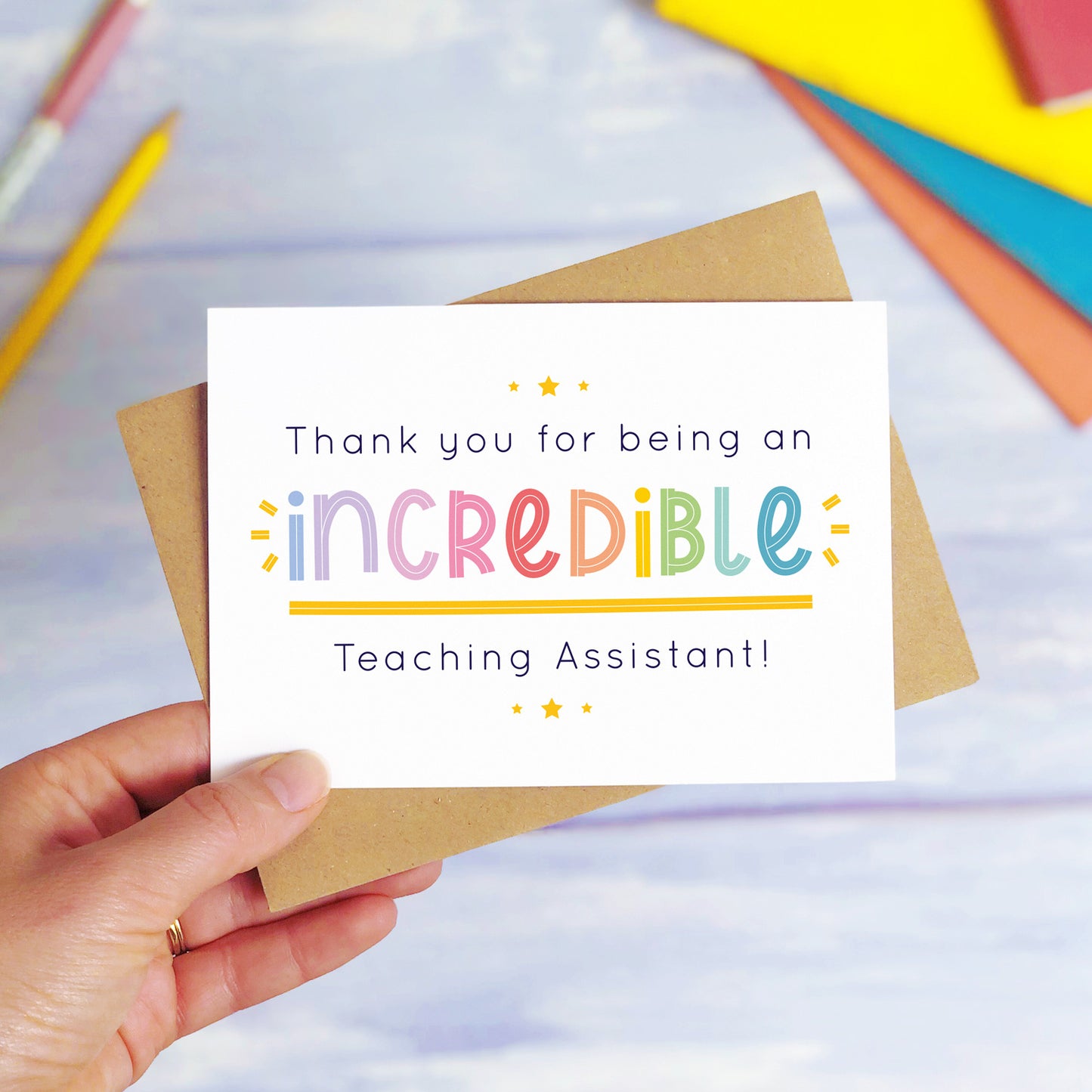 An incredible teaching assistant thank you card being held over a blue background with colourful text books and two pens. The card is sat on top of its kraft brown envelope. This is the teaching assistant card with the rainbow text colour option.