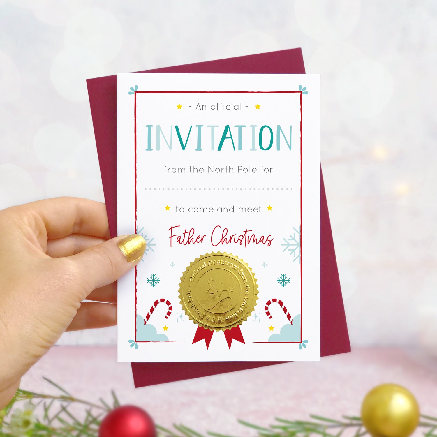 An invitation certificate to visit Santa (or Father Christmas) photographed in hand in front of a grey and dusty pink background with foliage and baubles in the foreground. The card has a blank space for you to write a childs name. It also features a shiny gold seal with the face of a santa character.