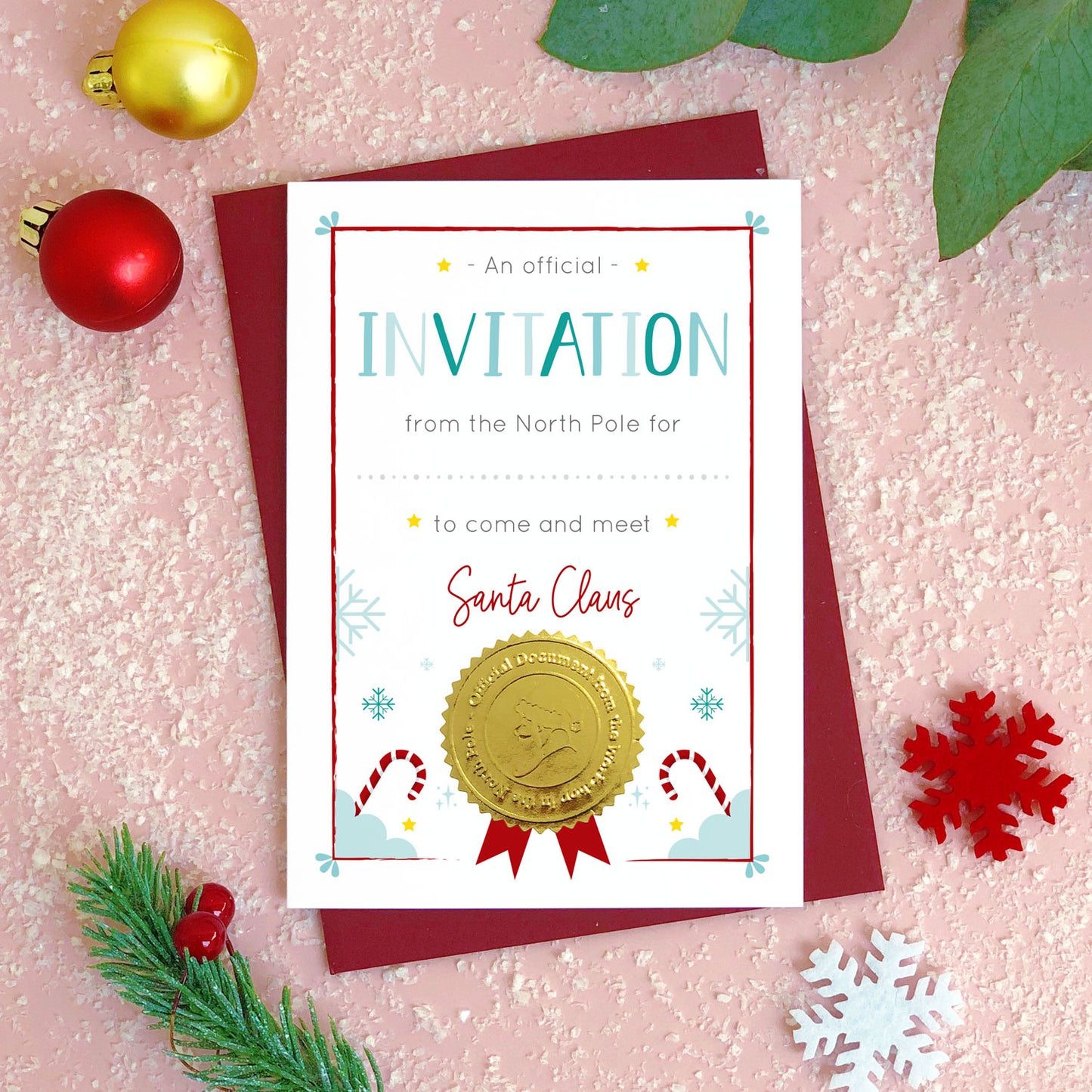 An invitation certificate to visit Santa (or Father Christmas) photographed flat lay style on top of a pink background covered in ‘snow’, pieces of foliage, snowflakes and a bauble. The card has a blank space for you to hand write a childs name. It also features a shiny gold seal with the face of a santa character.