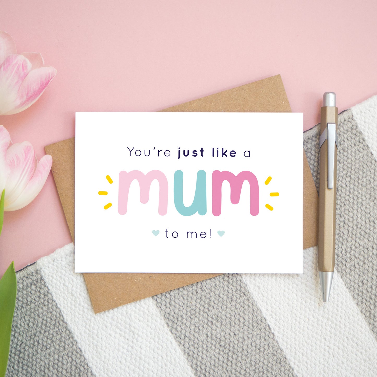 A flatlay set up of the pink and blue version of the 'you're just like a mum to me' card. It is landscape in orientation and is photographed next to a pen and pink tulips for scale.