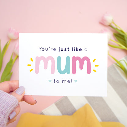 You're just like a mum to be me card in pink and blue being held in the left hand over a pink, white and grey background with tulips.