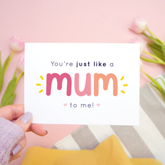 You're just like a mum to be me card in pink and peach being held in the left hand over a pink, white and grey background with tulips.
