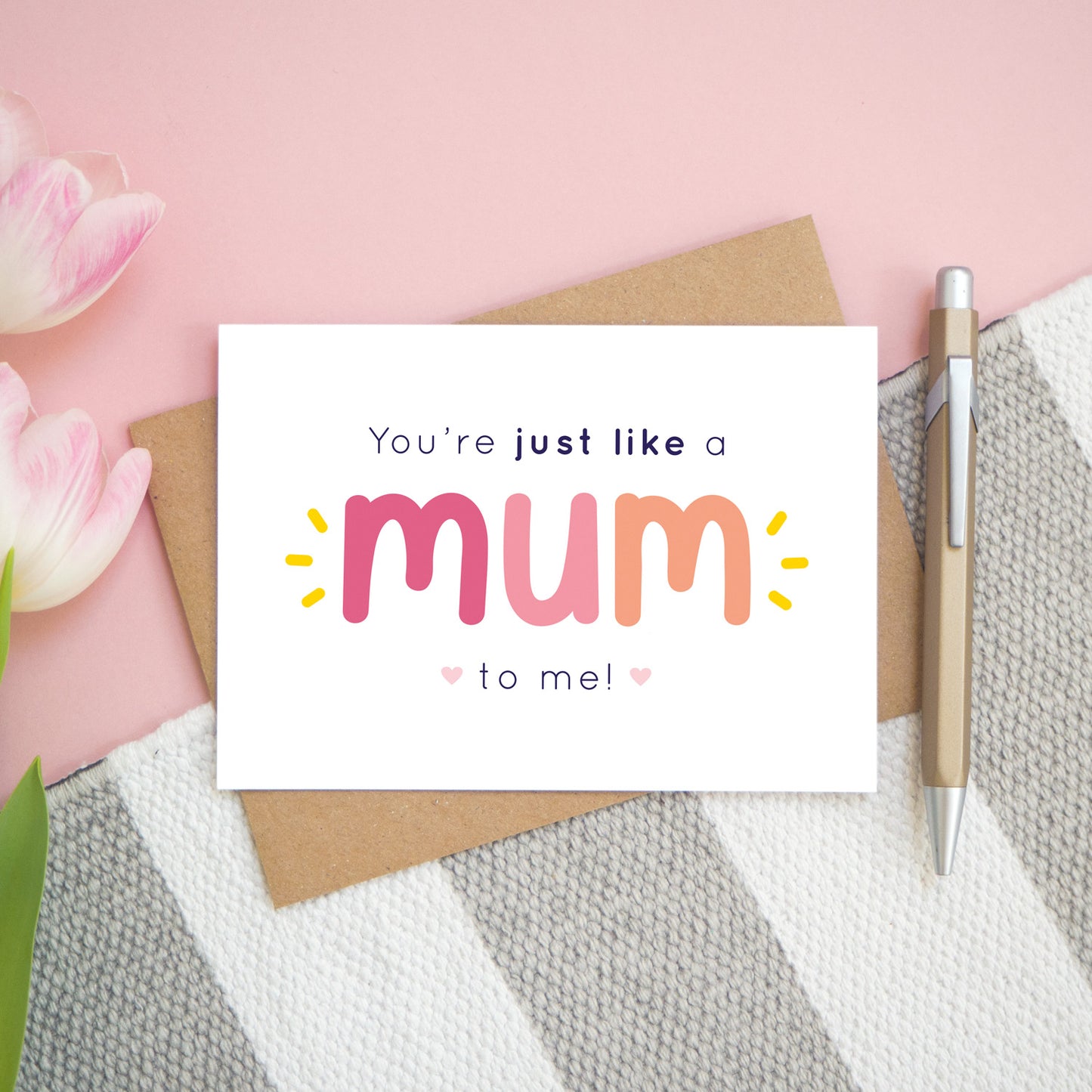 A flatlay set up of the pink and peach version of the 'you're just like a mum to me' card. It is landscape in orientation and is photographed next to a pen and pink tulips for scale.
