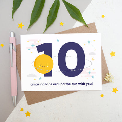 Laps around the sun anniversary card featuring a sun orbiting the number 10 surrounded by stars. Flat lay image shot on a white and grey background with some foliage, yellow stars and a pink pen.