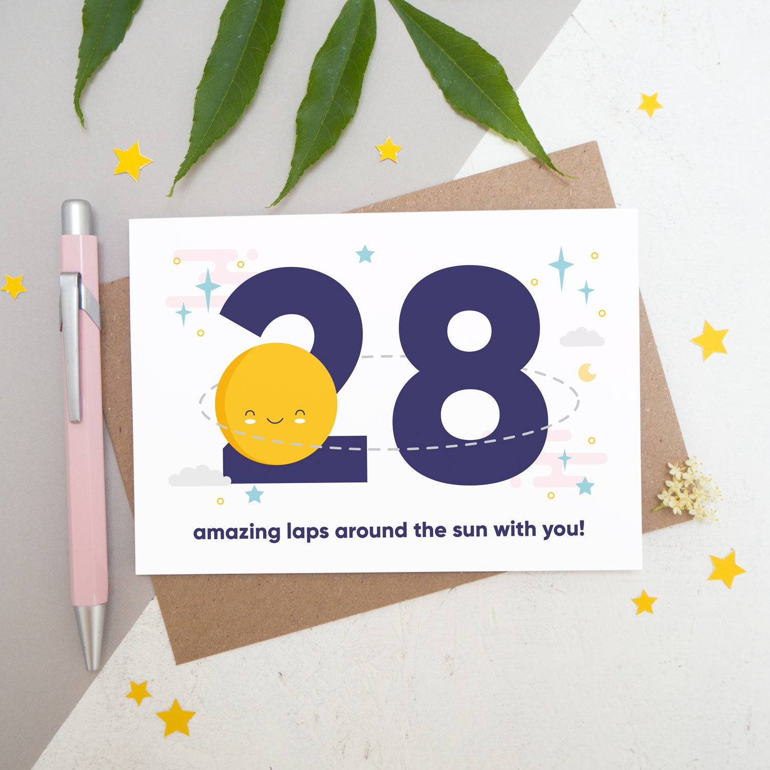Laps around the sun anniversary card featuring a sun orbiting the number 28 surrounded by stars. Flat lay image shot on a white and grey background with some foliage, yellow stars and a pink pen.