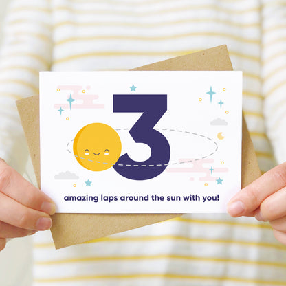 Laps around the sun anniversary card featuring a sun orbiting the number 3 surrounded by stars. Shot as a lifestyle image with someone in a white and yellow striped jumper holding the card.