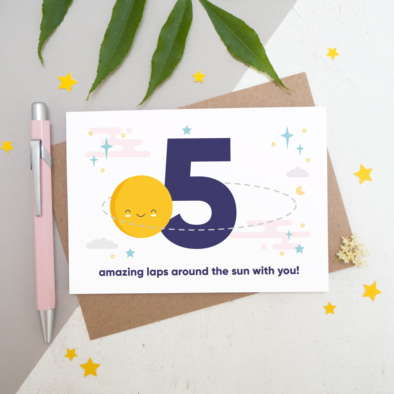 Laps around the sun anniversary card featuring a sun orbiting the number 5 surrounded by stars. Flat lay image shot on a white and grey background with some foliage, yellow stars and a pink pen.