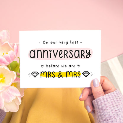 A last anniversary or Valentine’s card photographed on a pink background with pink tulip flowers, a grey and white stripe rug and yellow fabric. This image shows the last anniversary option with the Mrs & Mrs wording. The text is black and there are pops of yellow and pink behind key words.