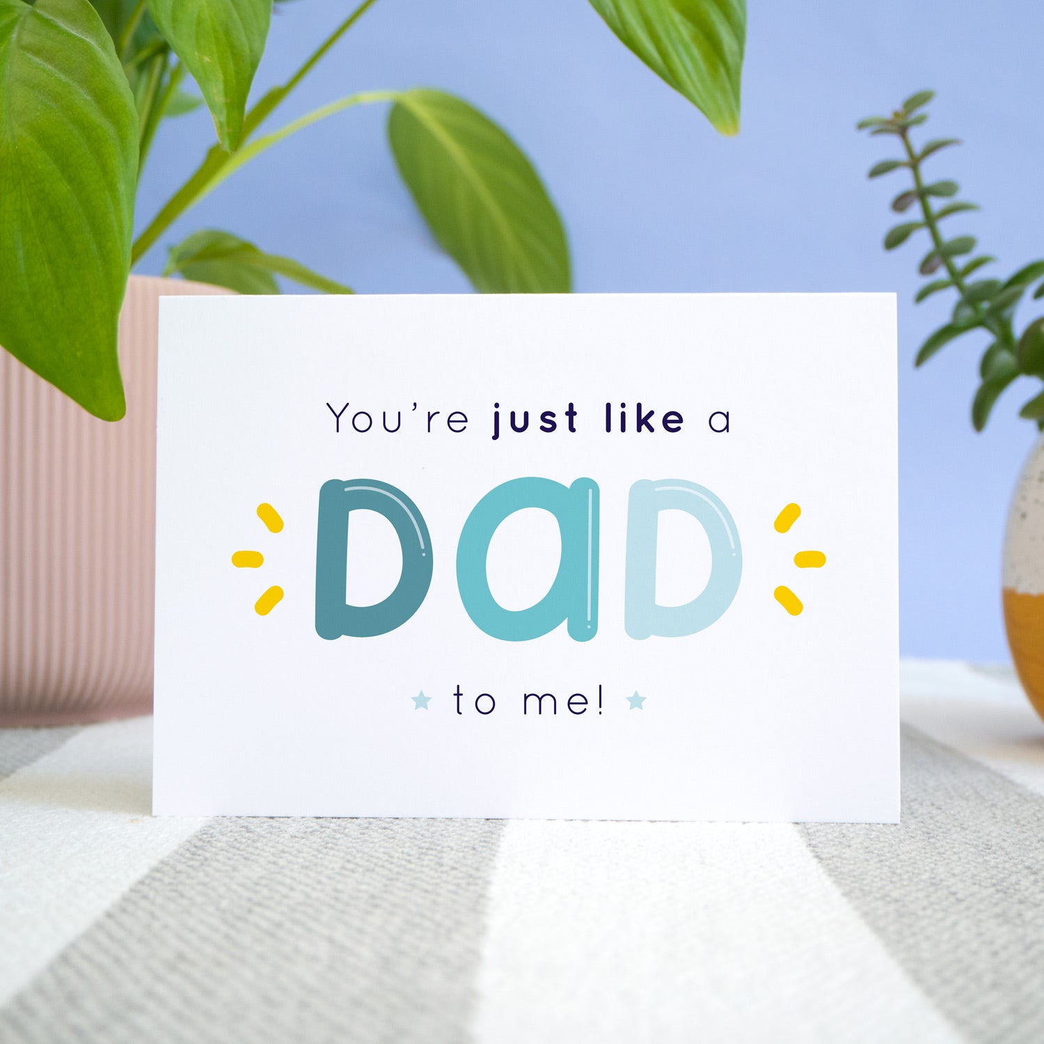 A 'like a dad to me' card shot on a stripy rug with a blue background with potted plants. The card shows the text 'you're just like a dad to me!' in varying tones of blue and navy with yellow flicks.