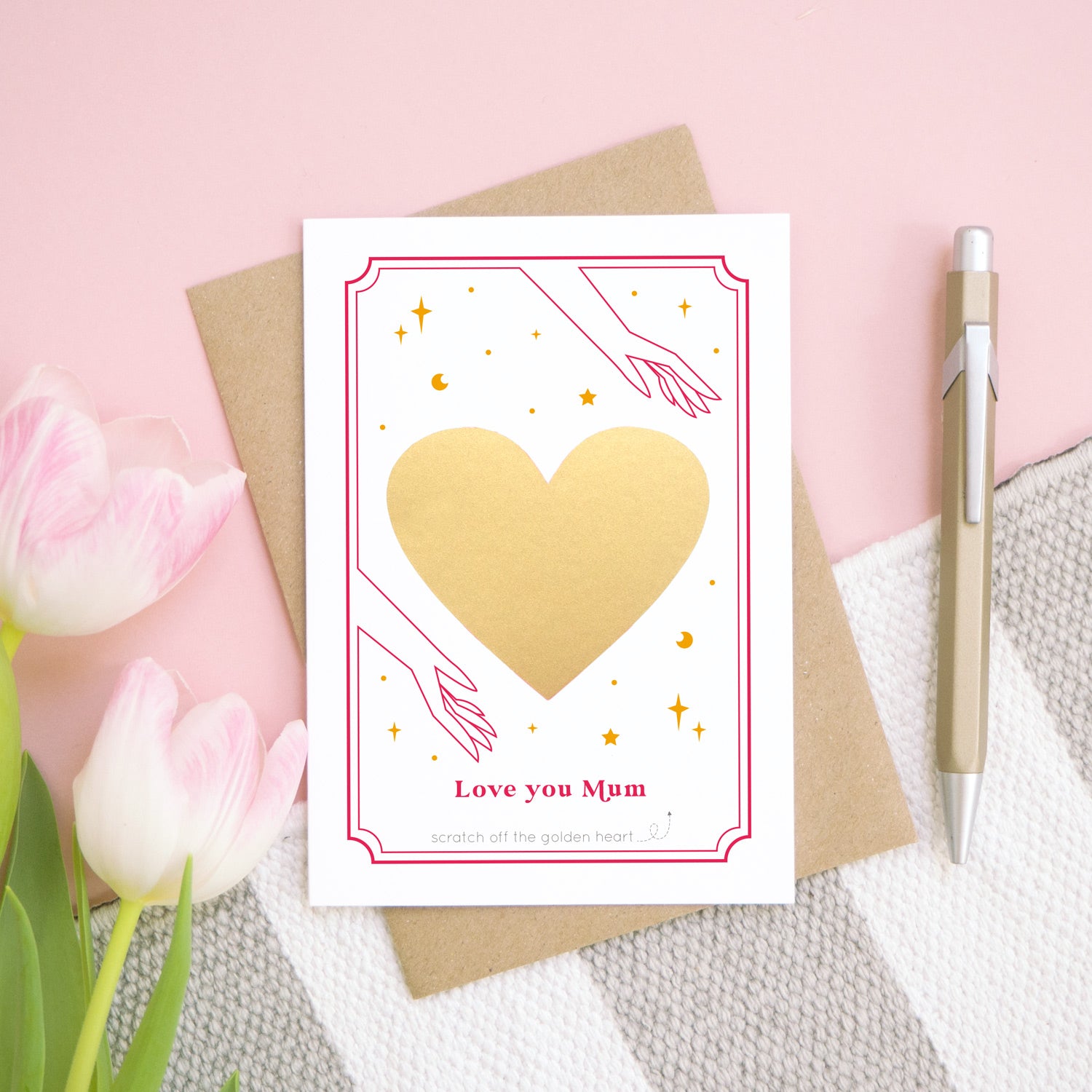 A personalised 'love you...' card before the gold heart has been scratched off. It has been shot on a pink background, with tulips and a pen for scale.
