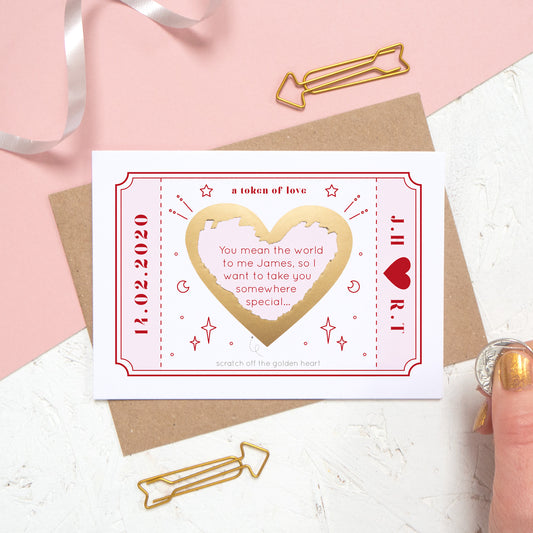 The personalised Love Token Scratch card with the printed message scratched off. Shot on a pink and white background with a hand in the corner.