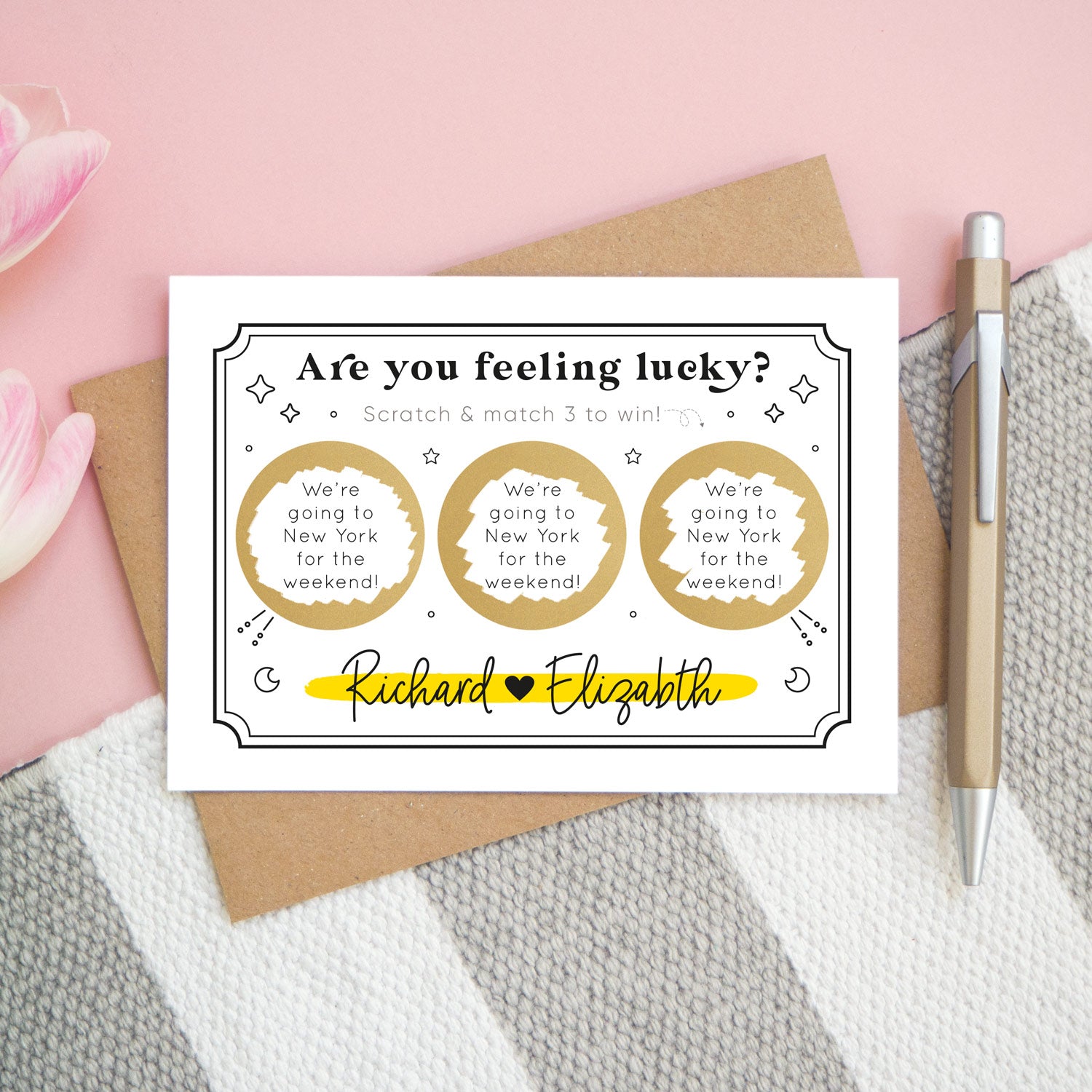 A personalised feeling lucky scratch card photographed on a pink and grey & white striped background with pink tulips poking in the side and a gold pen. The landscape scratch card sits on a kraft brown envelope. This card shows a celestial token style illustration in the black and yellow colour scheme with 3 golden circles which have been scratched off to reveal the winning message!