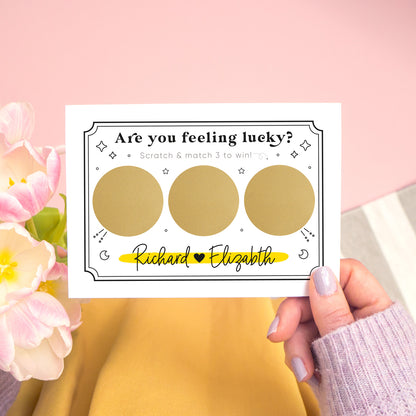 A personalised feeling lucky scratch card photographed on a pink and grey & white striped background with pink tulips and yellow fabric. A hand holds the landscape scratch card. This card shows a celestial token style illustration in the black and yellow colour scheme with 3 golden circles which have not yet been scratched off!