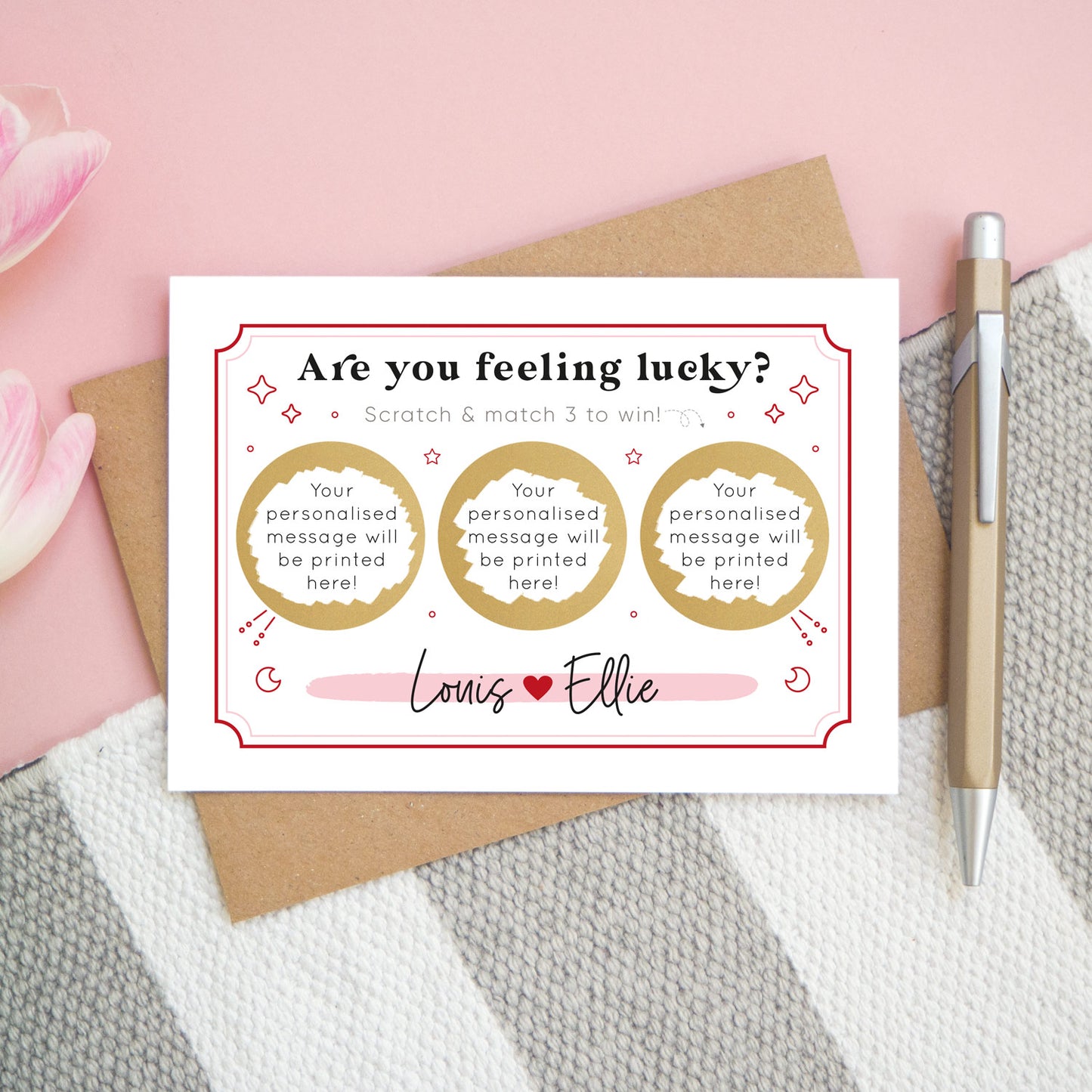 A personalised feeling lucky scratch card photographed on a pink and grey & white striped background with pink tulips poking in the side and a gold pen. The landscape scratch card sits on a kraft brown envelope. This card shows a celestial token style illustration in the red and pink colour scheme with 3 golden circles which have been scratched off to reveal the winning message!