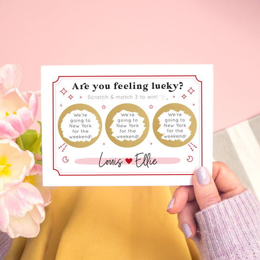 A personalised feeling lucky scratch card photographed on a pink and grey & white striped background with pink tulips and yellow fabric. A hand holds the landscape scratch card. This card shows a celestial token style illustration in the red and pink colour scheme with 3 golden circles which have been scratched off to reveal the winning message!