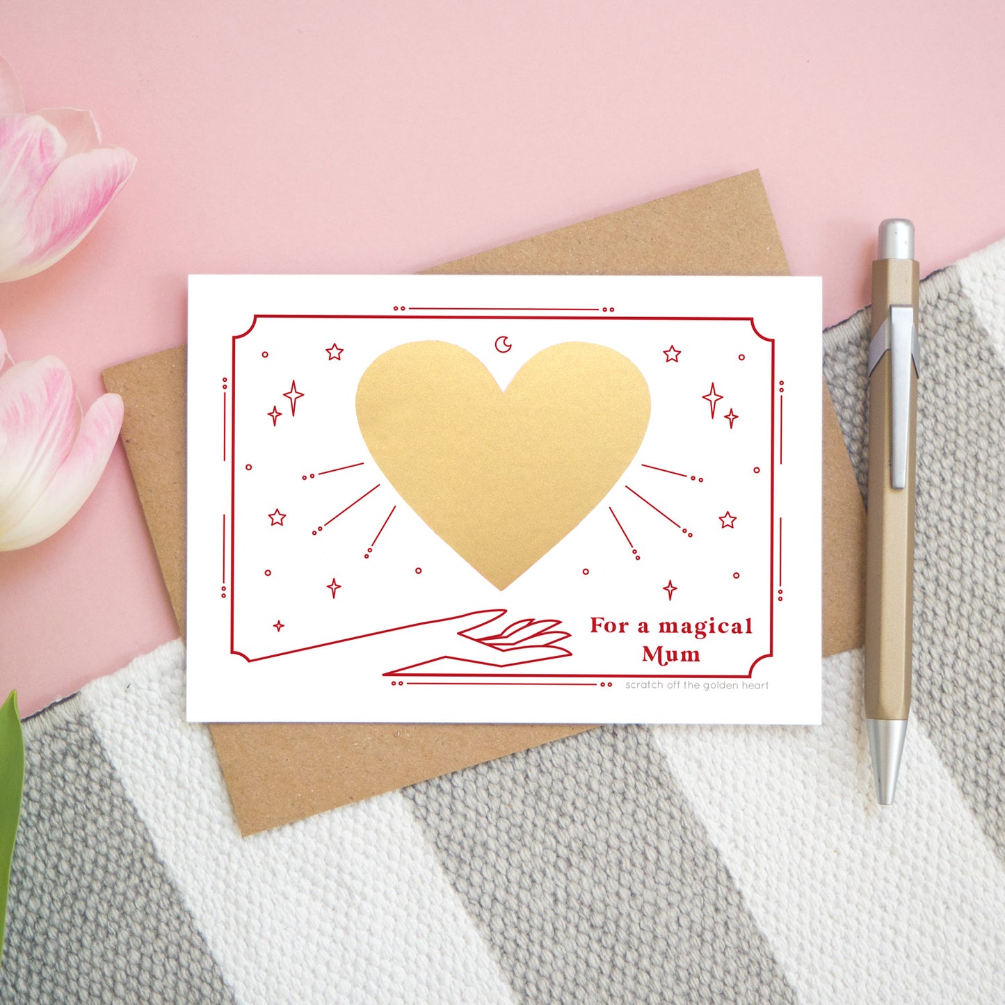 A personalised 'magical mum' scratch card before the gold heart is scratch off. Card is photographed from above on a pink, white and grey background with a pen for scale.