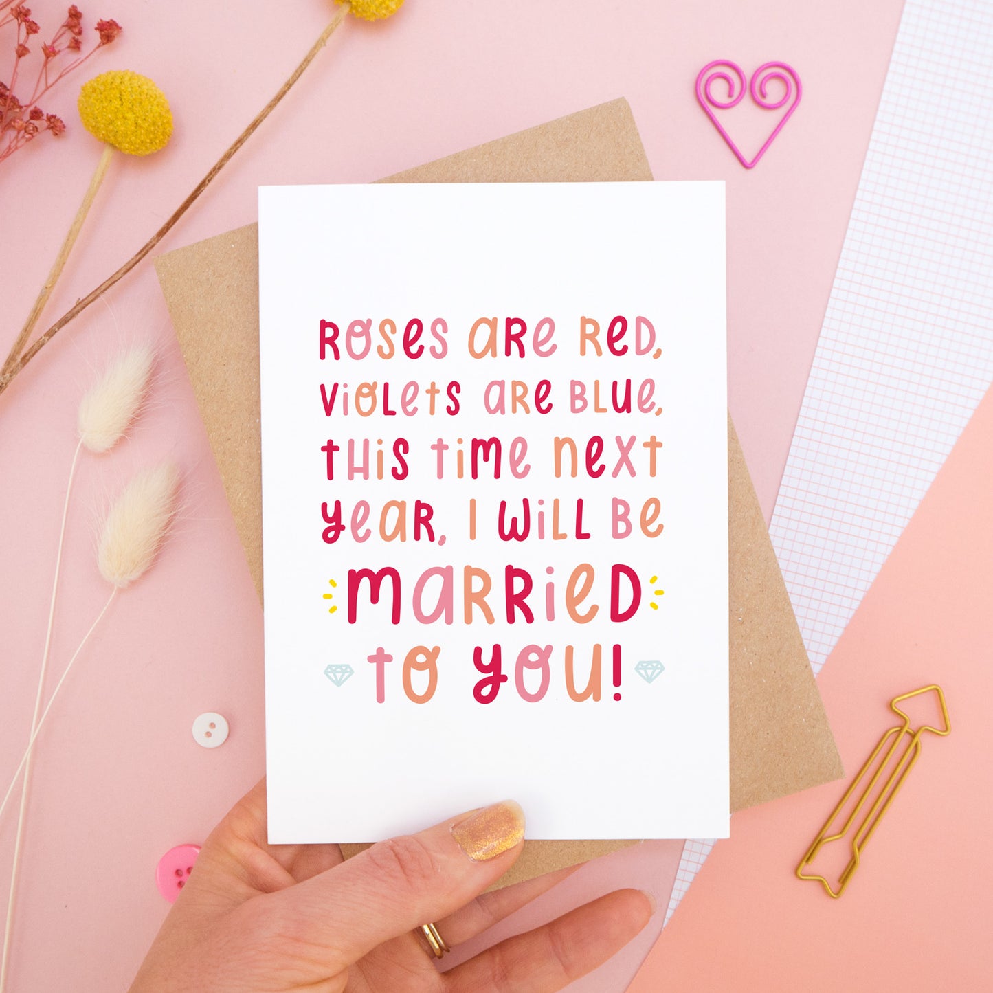 The 'this time next year' card photographed on a pink background with dried flowers, buttons and paper clips as props. The card itself is being held above the scene.
