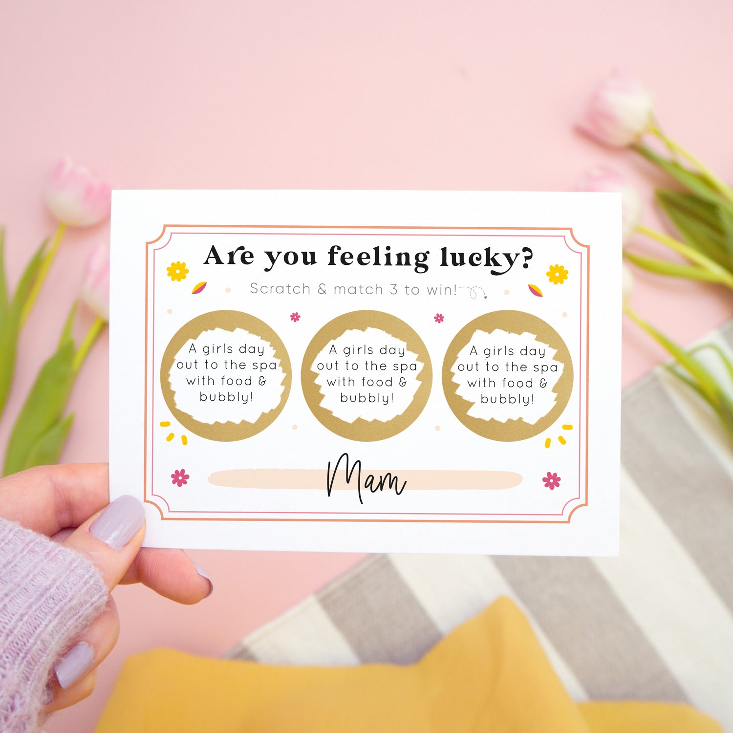 A personalised floral match 3 to win style scratch card being held over a pink, grey and white background with tulips scattered around the edges. The card has had all 3 golden scratch panels scratched off to reveal the 3 matching winning messages! This card is the pink and peach version.